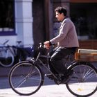 Ein Mann fährt ein Fahrrad mit einer orangefarbenen Kiste hinten. Hintergrund unscharf, andere Fahrräder sichtbar.