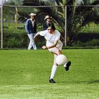 Eine Frau in weißem Trikot schießt einen Fußball auf einem grünen Spielfeld, im Hintergrund stehen zwei Männer.