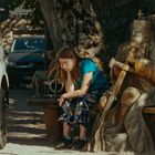 Ein Mädchen sitzt auf einer Bank neben einer großen Holzskulptur eines Königs mit Schwert und Krone unter einem Baum.