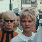 Eine Gruppe ernst blickender Menschen auf einer belebten Straße, Hintergrund mit Gebäuden und US-Flagge.