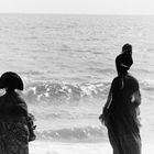 Keine sichtbaren Texte. Zwei Menschen in traditioneller Kleidung stehen am Strand und blicken aufs Meer.