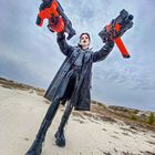 Eine Person in Ledermantel hält zwei große Spielzeugwaffen. Dramatischer Himmel und Sandboden im Hintergrund.