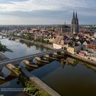 "Donaukanäle und Weinlände, links und rechts der Steinernen Brücke, bezeichnen historische und aktuelle Uferzonen zur Donau." Im Hintergrund eine Altstadt mit einer dominanten, gotischen Kathedrale und einer Flusslandschaft.