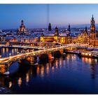 Abendansicht einer Stadt mit beleuchteter Brücke über einen Fluss, umgeben von historischen beleuchteten Gebäuden.