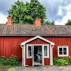 Ein rotes Holzhaus mit weißen Rahmen. Zwei Schornsteine und ein Fahrrad im Eingang. Bäume im Hintergrund, bewölkter Himmel.