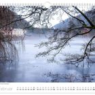 "Februar, Frostige Stimmung am Schlossteich." Ein zugefrorener Teich mit kahlen Bäumen und einem rosa Gebäude im Hintergrund.