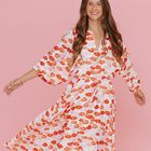 Keine Texte vorhanden. Eine lächelnde Frau in einem Kleid mit rotem Kussmundmuster vor rosa Hintergrund.