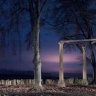 Text: „MACHT UND MYTHEN“. Zwei Bäume und ein Steinbogen bei Nacht, im Hintergrund ein violetter Himmel.
