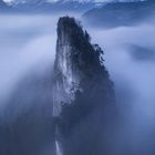 Ein hoher, bewaldeter Felsen ragt aus dichtem Nebel empor, dahinter schneebedeckte Berge.