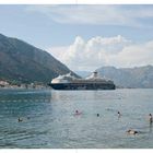 „Mein Schiff“ auf einem großen Kreuzfahrtschiff vor einer malerischen Bergkulisse; Menschen schwimmen im Meer.