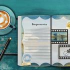Text: "Borgerservice", "Umzug". Aufgeschlagenes Notizbuch, daneben Stift und Tasse mit Latte Art auf Holztisch.