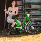 Februar-Kalenderblatt, Frau in weißem Kleid auf grünem Moped, Heugabel in der Hand, rustikaler Hintergrund.
