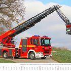 August: Ein Feuerwehrwagen mit ausgefahrener Drehleiter steht auf einem Feldweg, daneben ein großer, kahler Baum.