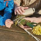 Ein Kind und ein Erwachsener binden Moos und eine gelbe Hyazinthe mit Schnur auf einem Holztisch zusammen.