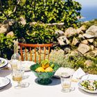 Tisch mit Zitrusschale, Gläsern und Tellern, darunter Blumendekor, im Freien, umgeben von Steinmauern und grüner Natur.