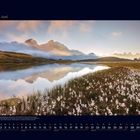 06 Juni. Ein Bergsee spiegelt majestätische Berge. Im Vordergrund blühende weiße Blumen unter einem klaren Himmel.
