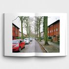 Straße mit geparkten Autos, links und rechts rote Gebäude. Bäume stehen am Straßenrand entlang.