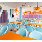 Keine Texte vorhanden. Moderne Kantine, bunte Tische, blaue Stühle, orange Lampen, pastellfarbene Wände.