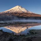 Ein schneebedeckter Vulkan spiegelt sich in einem kleinen See; die Landschaft ist karg und es ist früh am Morgen.
