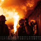Zahlen und Wochentage am unteren Rand; Feuerwehrleute in Schutzkleidung vor lodernden Flammen.
