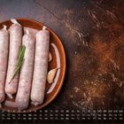 Kalender mit Monatstagen und numerischer "2". Vier rohe Würste und Rosmarin auf einem braunen Teller, daneben Knoblauch.