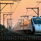 Text: "Solapur Jn" und eine Kalenderübersicht. Ein Zug fährt auf Gleisen bei Sonnenuntergang. Stromleitungen ziehen sich entlang.