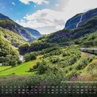 "Flamsbana, normalspurige Eisenbahnstrecke." Ein Zug fährt durch eine bergige, grüne Landschaft mit blauem Himmel.