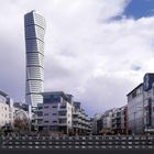Text: "Stadtbild Malmö 2". Ein modernes Gebäude mit verdrehter Form vor einem wolkigen Himmel, umgeben von weiteren Gebäuden.