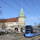 Straßenbahnlinie 2, blaues Fahrzeug, fährt vorbei an Gebäude mit Turm und roten Ziegeldächern unter klarem Himmel.