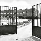 Eine offene Gittertür führt zu einem Gebäude mit industriellem Aussehen, umgeben von einer Grünfläche.