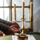 Eine Hand rührt in einem kleinen Kupfertopf auf einem grün gedeckten Tisch, im Hintergrund brennen Kerzen.