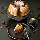 Ein Gugelhupf mit hellem Zuckerguss und Kirschen; eine Scheibe auf einem Teller mit einer Espressotasse daneben.