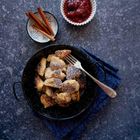 Kein erkennbarer Text. Gezeigt wird gebratener Teig in einer Pfanne, Zimtstangen und rote Sauce in einer Schale.