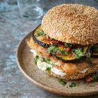 Ein Sesam-Bagel mit gegrillten Auberginen, frischen Kräutern und einer Creme auf einem rustikalen Teller.