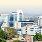 Text: "WINDHOEK, KHOMAS-HOCHLAND UND KALAHARI". 

Stadtansicht mit modernen Gebäuden und Bäumen im Vordergrund.