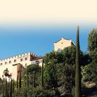 Text: "MERAN UND UMGEBUNG". Schlossartige Gebäude vor Pinien und Zypressen unter klarem blauem Himmel.