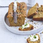 Ein Laib Brot auf einem Teller, bestreut mit Kürbiskernen. Ein Messer liegt darauf. Brotstücke mit Kräutercreme daneben.