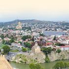 Text: "TBILISI". 
Stadtbild mit Fluss, Hügeln, vielen Gebäuden, Kirchen und einer Festung. Ruhige, grüne Umgebung.