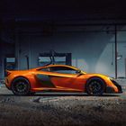 Ein leuchtend orangefarbener Sportwagen steht in einer verlassenen, leicht verfallenen Fabrikumgebung auf einem unebenen Boden.