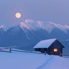 Verschneite Hütte mit leuchtendem Fenster, Mond über Bergkette, ruhige Abendstimmung.