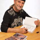 Ein älterer Mann signiert Bücher. Er trägt ein schwarzes Kopftuch und ein verziertes Shirt. Bücher sind vor ihm gestapelt.