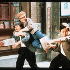 Mehrere chinesische Schriftzeichen auf Schildern. Eine dynamische Szene: Drei Männer sind in einer Auseinandersetzung verwickelt.