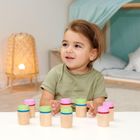 Ein Kleinkind sitzt an einem Tisch, umgeben von bunten Holzspielzeugen. Im Hintergrund steht eine Lampe und ein Bett mit Kissen.