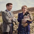 Zwei Personen stehen vor einer Steinmauer in einer ländlichen Landschaft. Eine Frau hält einen flauschigen Hund im Arm.