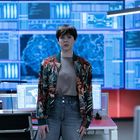 Mehrere Bildschirme zeigen technische Diagramme. Eine Person steht vor einem Schreibtisch, trägt eine bunte Jacke.