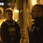 Text auf Uniform: "POLICE". Drei Personen im Gespräch auf einer nächtlichen, gelb beleuchteten Straße.
