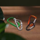 Grüne, silberne, orange und schwarze Karabiner liegen auf einem Holztisch, teils ineinander verschlungen. Links Pflanzenblätter.