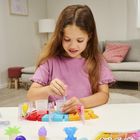Ein Mädchen in einem lila T-Shirt spielt am Tisch mit bunten Plastikformen und einer chemischen Spielzeuglösung. Im Hintergrund sind ein Sofa, Kissen und Pflanzen zu sehen.