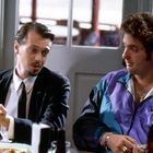 Zwei Männer sitzen nebeneinander. Links trägt ein Mann einen Anzug, rechts trägt einer eine lila Jacke mit einem Nike-Logo.