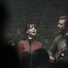 Zwei Personen singen in einem dunklen Raum vor einem alten Mikrofon. Eine Gitarre ist im Hintergrund zu sehen.
